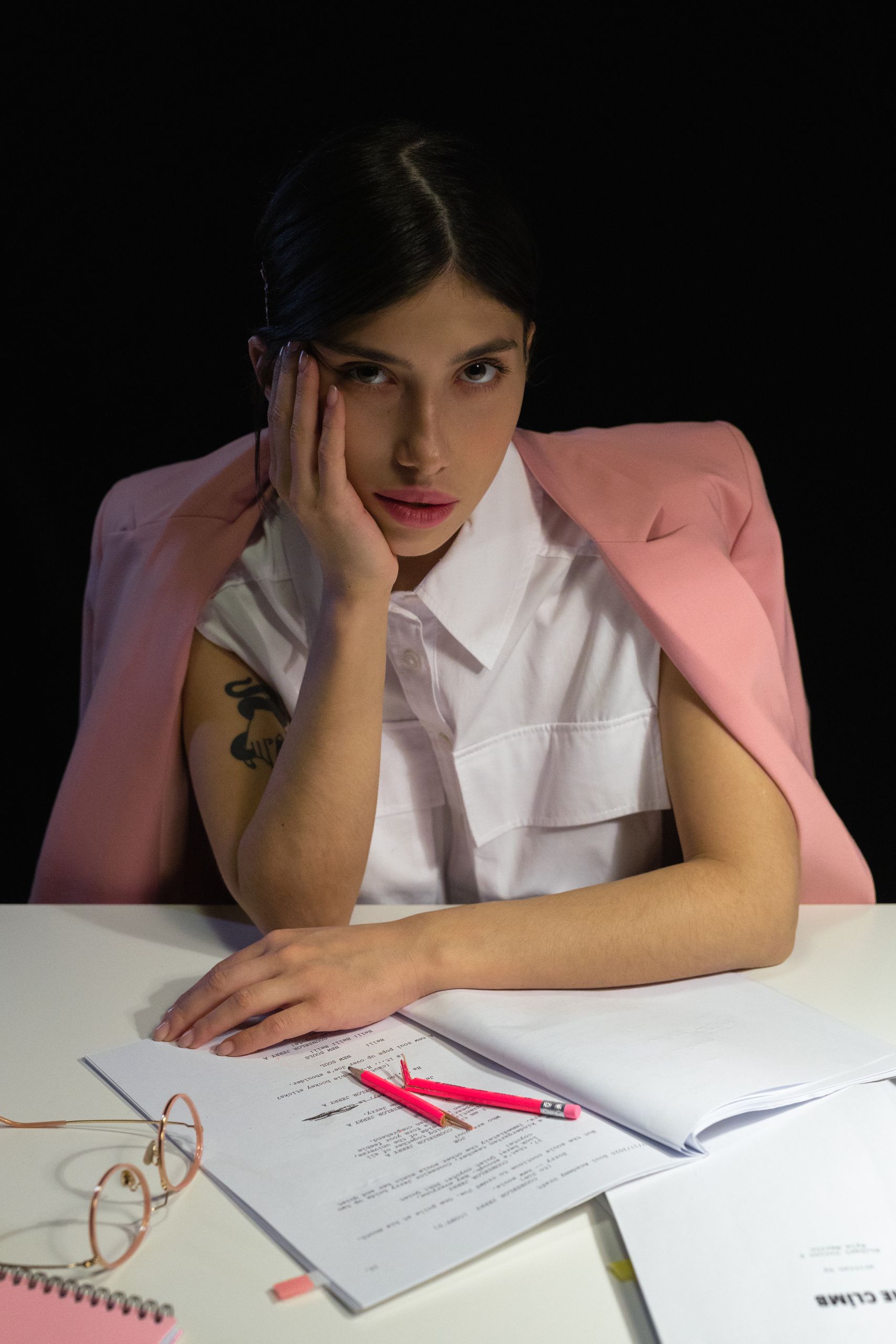 Young woman in a pink blazer sitting at desk with papers, looking tired and thoughtful.
